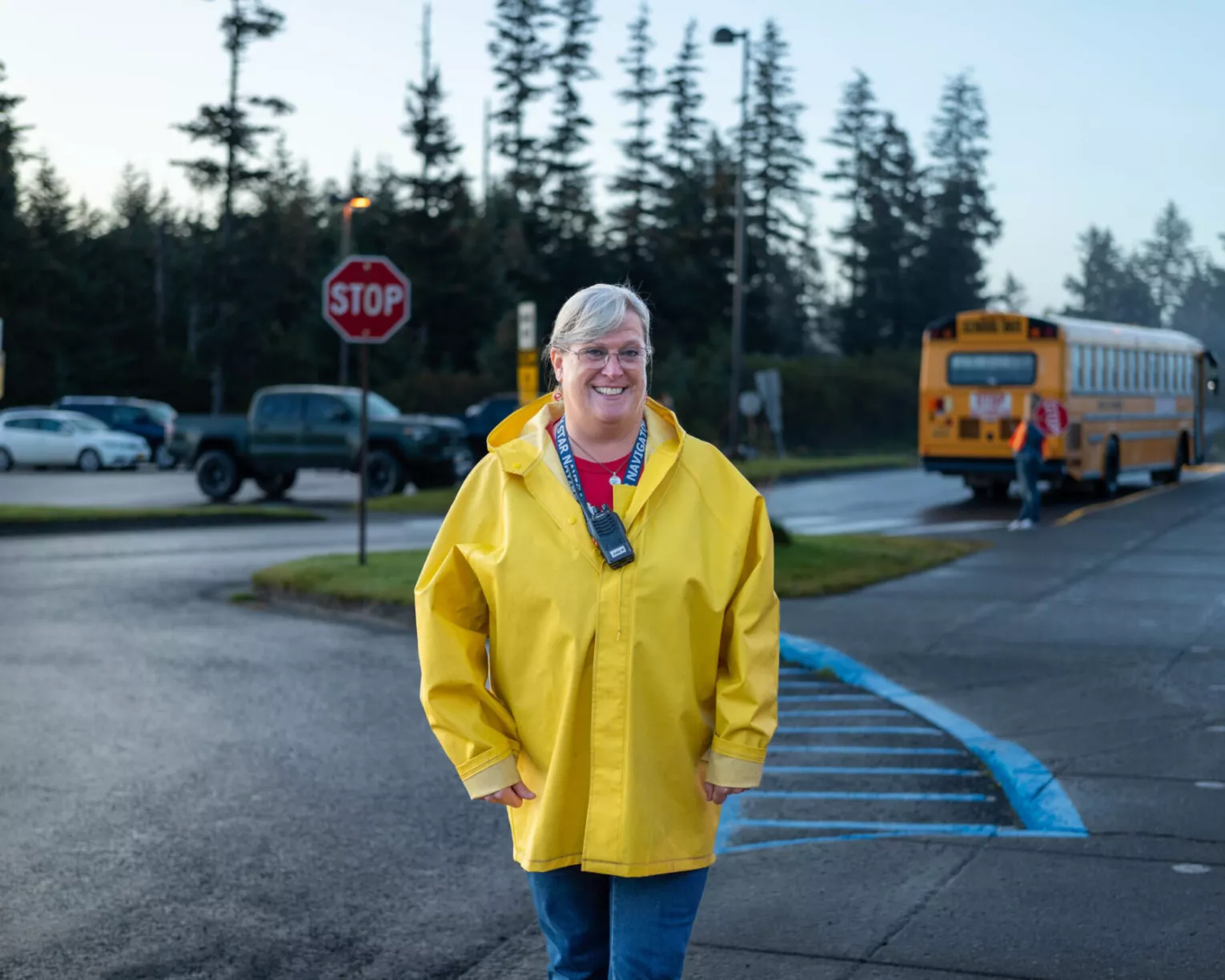 Julia Walker stands in front of the crosswalk at the school where she works in a bright yellow jacket. She is posing with a big smile. Behind her you can see pine trees, a stop sign, and a yellow school bus.