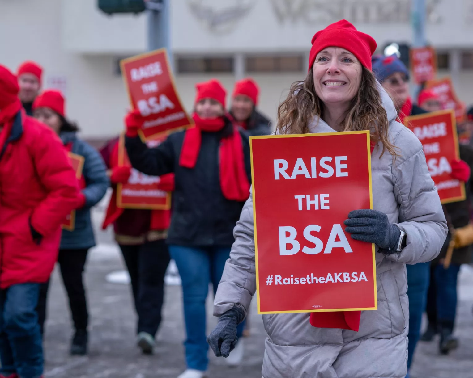 A photo from the Raise the BSA rally focused on a woman in the crowd of marching members. She is wearing a grey coat, red hat, and red scarf with a smile as she looks to the horizon with a hopeful look in her eyes. A sign that says "Raise the BSA" is in one of her gloved hands.