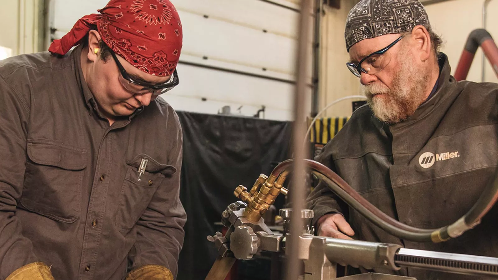 Robert Frasher works closely with a student in his classroom. They are both wearing bandanas and safety goggles as they concentrate on a task using welding equipment.