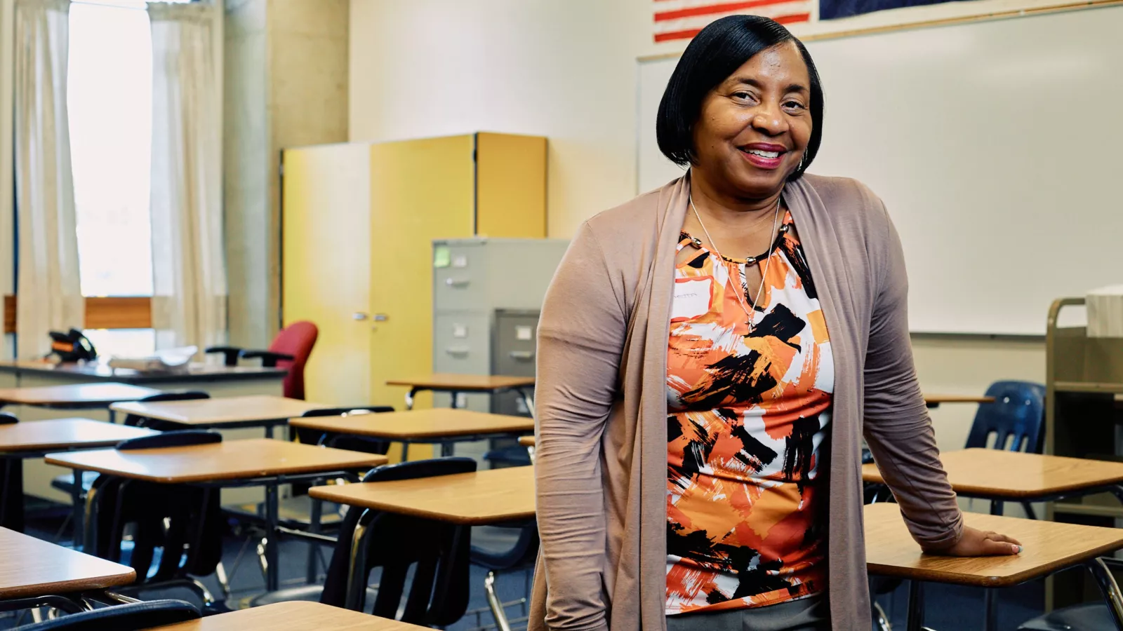 An African American teacher poses for a picture while leaning on a desk in an empty classroom.