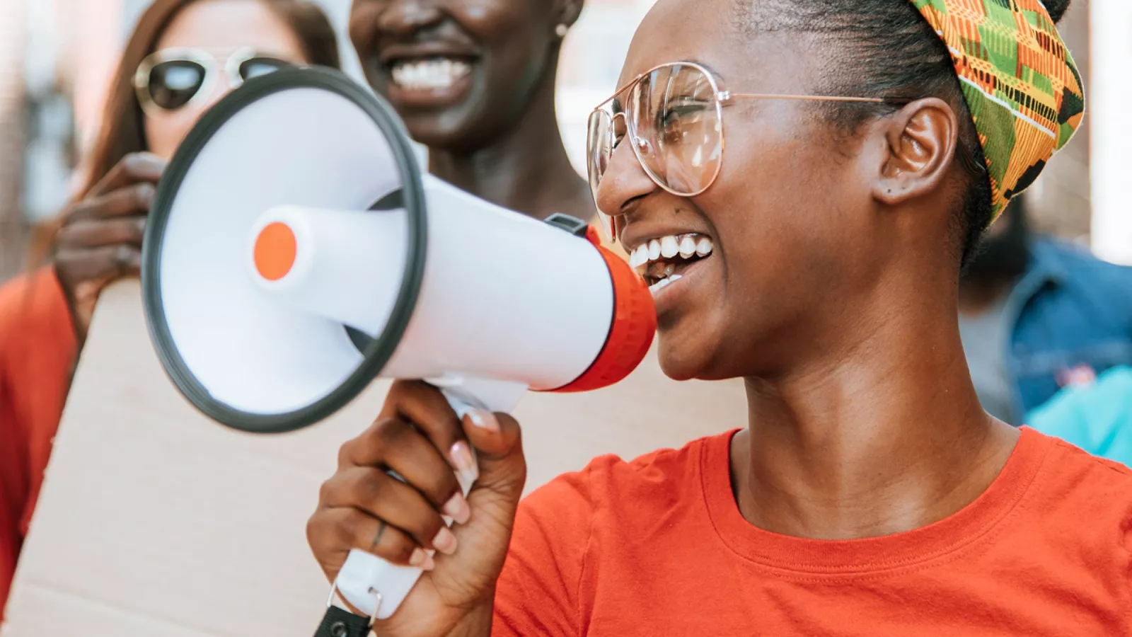 A young black woman holds a megaphone. She appears to be laughing. There are people behind her out of focus.