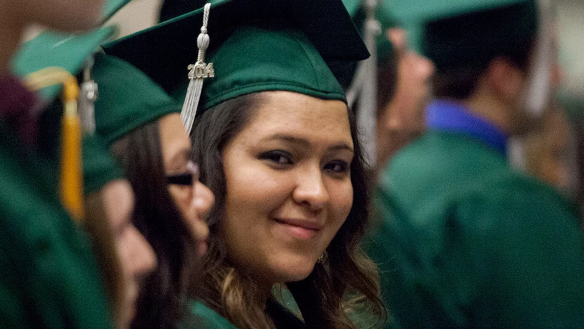 An aspiring educator at their graduation smiles softly at the camera. They are wearing a green cap with a 2014 graduate emblem.
