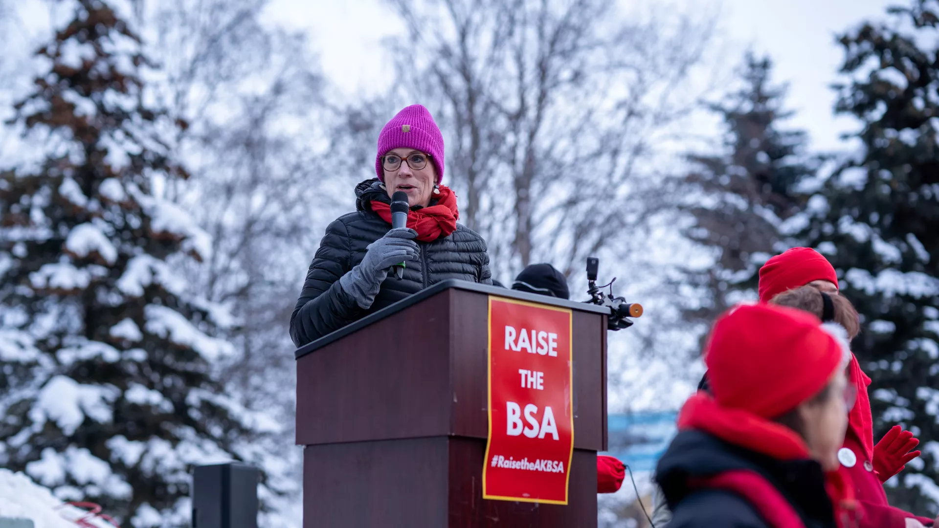 A woman in a pink hat speaks into a microphone at the podium at a rally in Alaska.