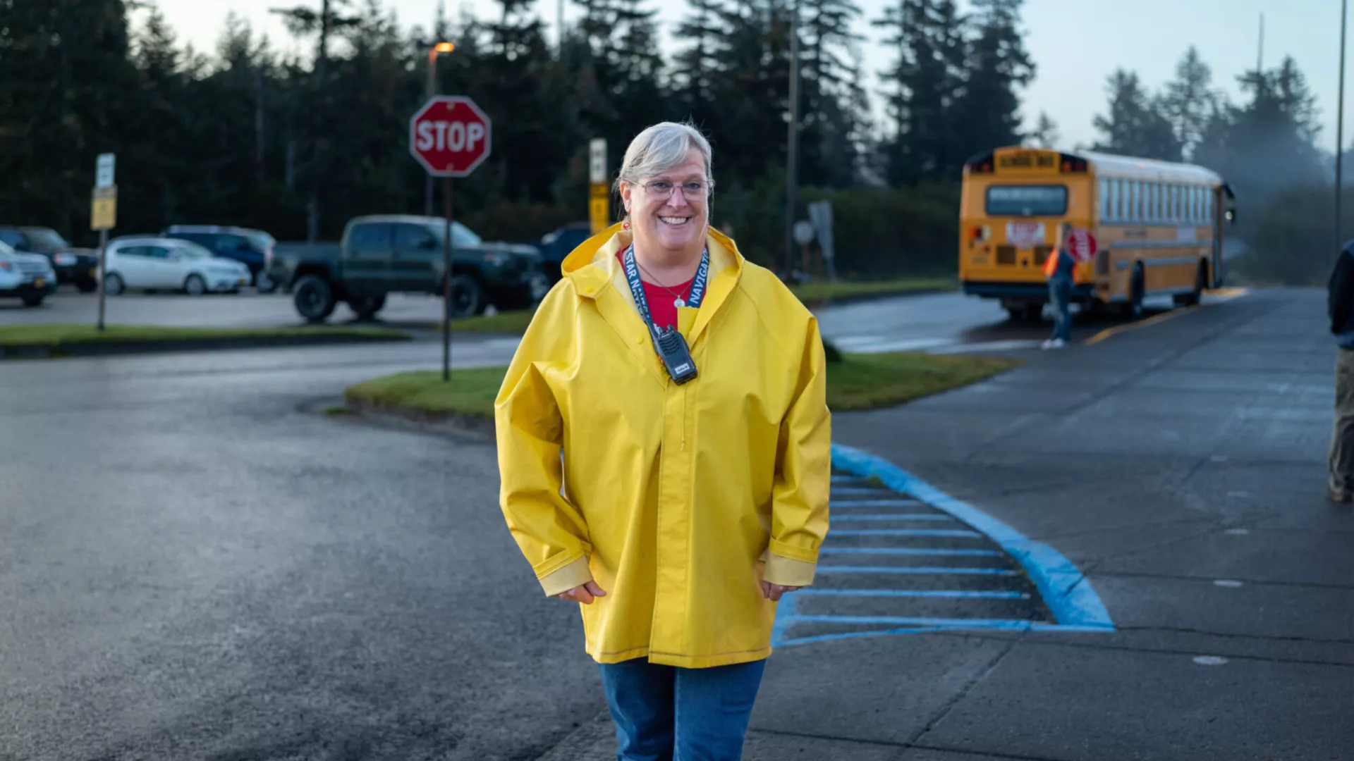 Julia Walker stands in front of a crosswalk wearing a bright yellow coat. In the background, there is a bus, a stop sign, a parking lot, and a view of pine trees.