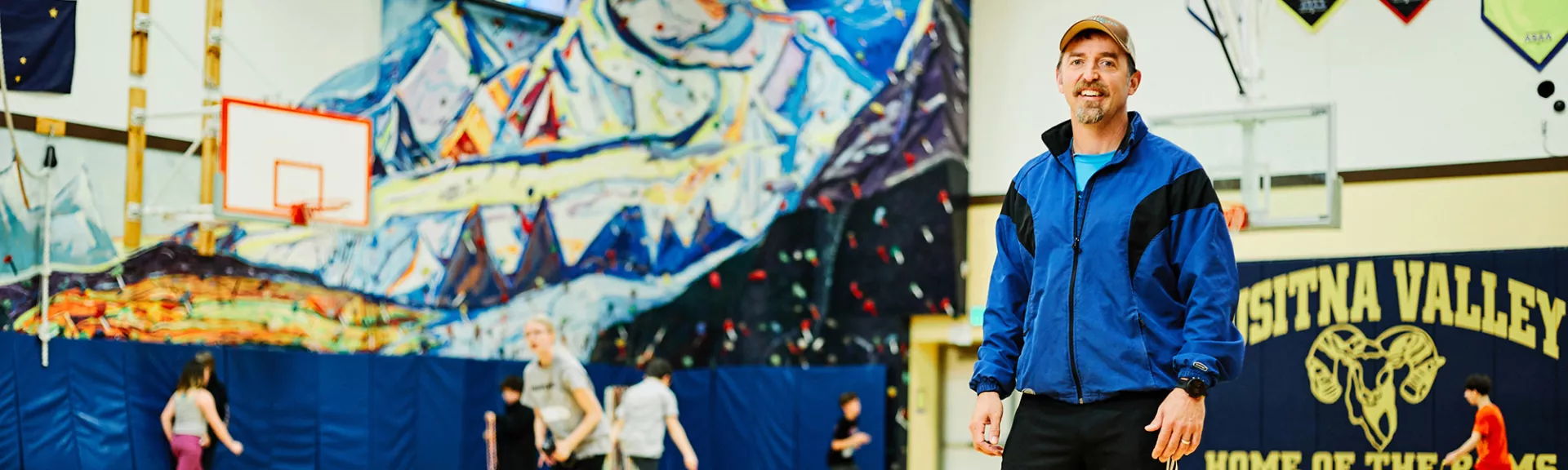 A gym teacher poses in a gymnasium with students playing basketball in the background.