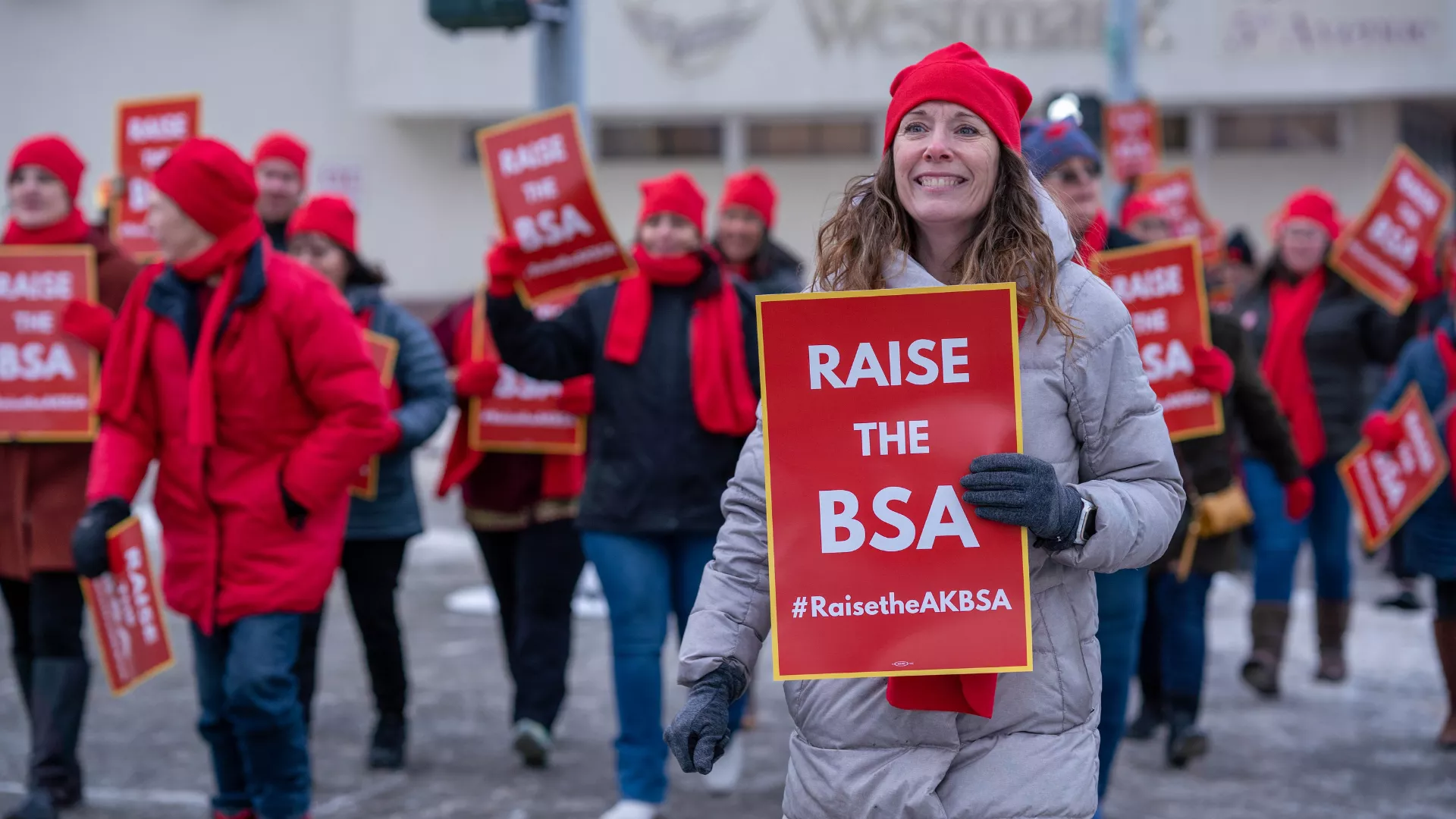 A photo from the Raise the BSA rally focused on a woman in the crowd of marching members. She is wearing a grey coat, red hat, and red scarf with a smile as she looks to the horizon with a hopeful look in her eyes.  A sign that says "Raise the BSA" is in one of her gloved hands.