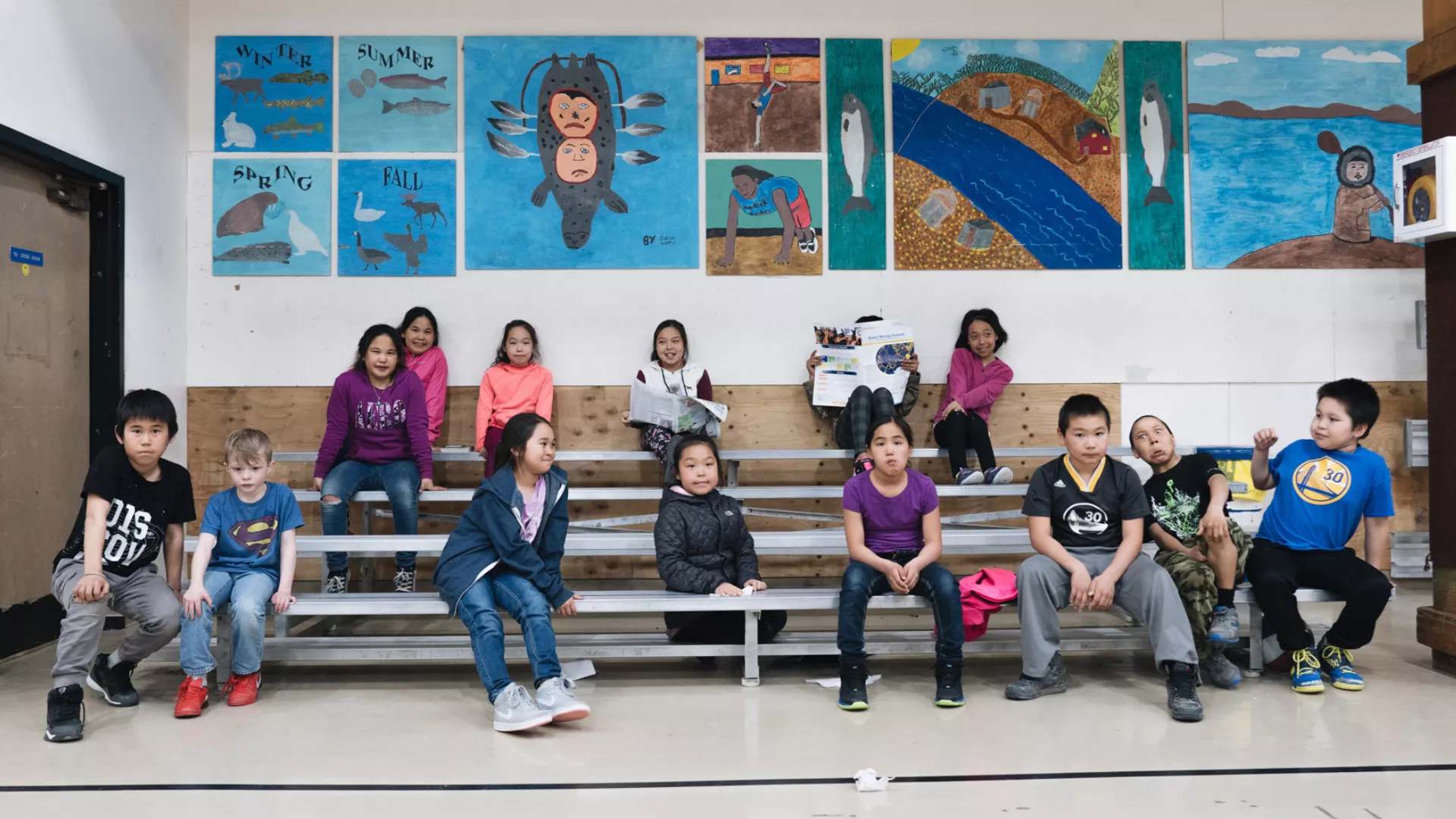 A group of Alaska native children sit in the bleachers at a school gymnasium with native art in the background.