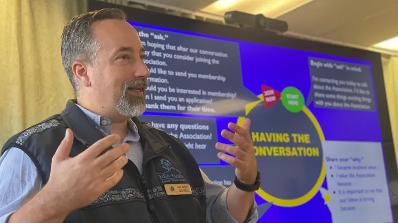 A photo of a light-skinned gentleman with salt-and-pepper hair and a dark-grey NEA-Alaska vest engaged a group of people offscreen in rigorous discussion