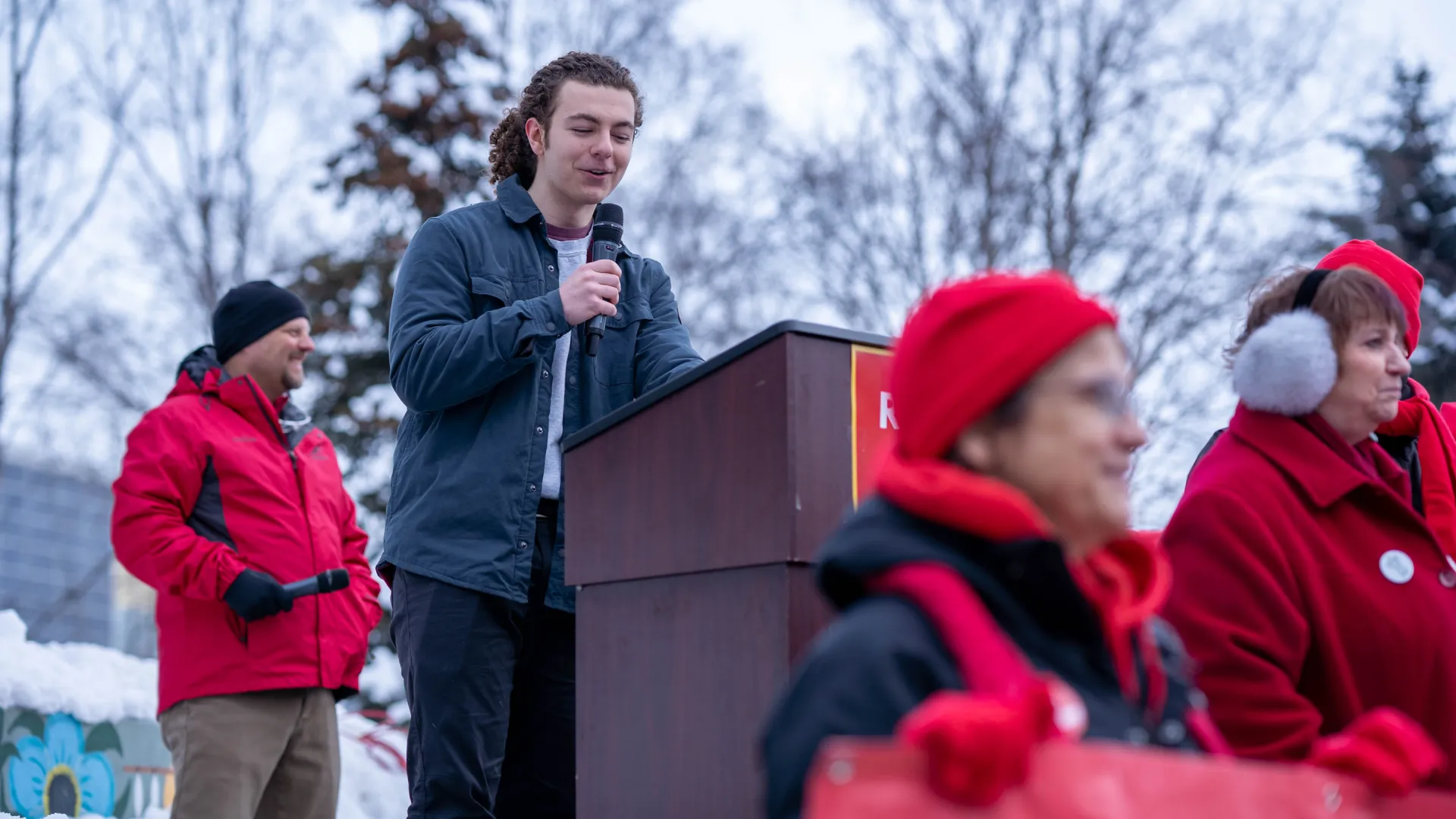 A young man stands outside at a podium with a microphone, addressing people wearing red