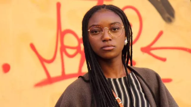 A Black girl stands in front of a yellow-gold wall with a determined expression