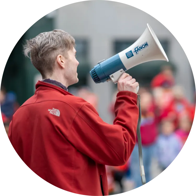 An image of a person with light skin and short, light-brown hair in a red jacket holding a white and blue megaphone with their back to the camera