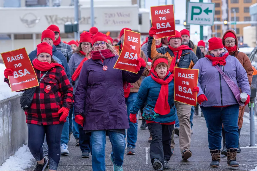 NEA-Alaska members hold red signs that say "Raise the BSA #RaisetheAKBSA" as they march in a rally. The members are bundled up for winter weather and wearing red accessories and buttons.