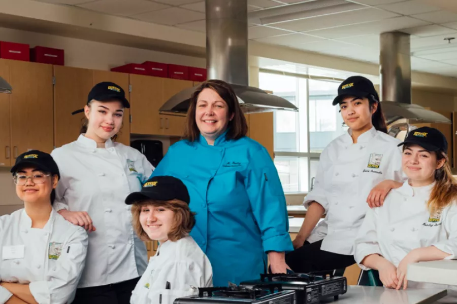 Melinda Dooley and her culinary students pose for a picture in their classroom. She is wearing a blue chef's coat, while they are wearing white chef's coats and black baseball caps.