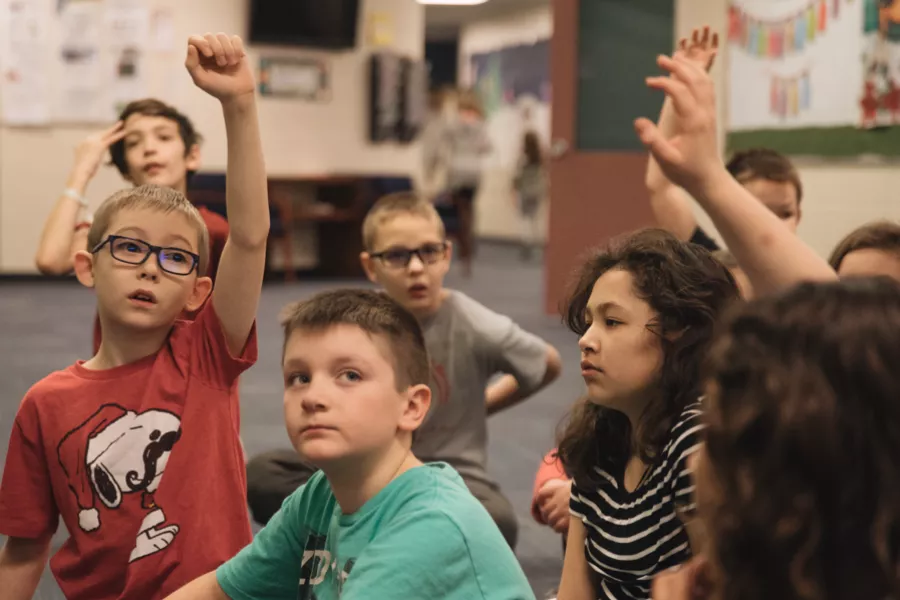 students in an elementary classroom raise their hands with eager expressions